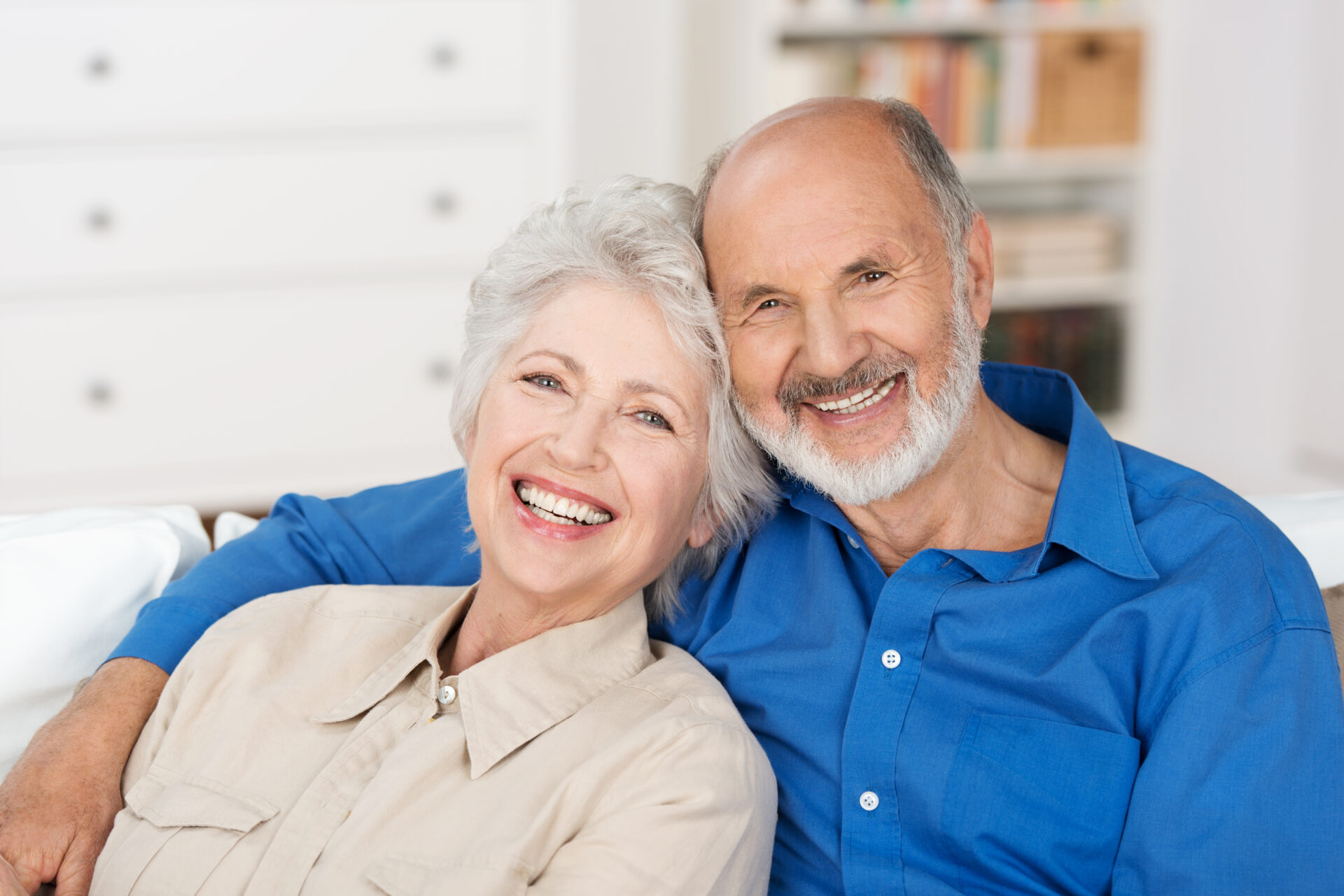 Romantic senior couple sitting close together on a sofa in the house smiling happily at the camera